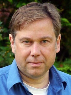 Matthias Bode mit ernstem Blick und blauem Hemd vor natürlichem Hintergrund.